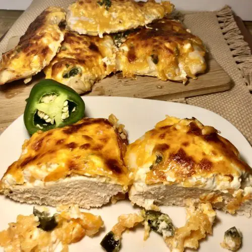 jalapeno popper smothered chicken breast with cream cheese and cheddar cheese. chicken breast is sliced in half on white plate with cutting board full of jalapeno popper smothered chicken breast in the background