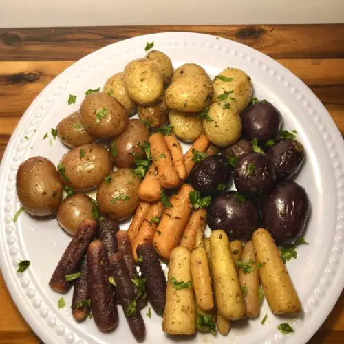 roasted rainbow carrots and baby potatoes plated on white plate against wooden cutting board. fresh cut parsley used as garnish over the top.