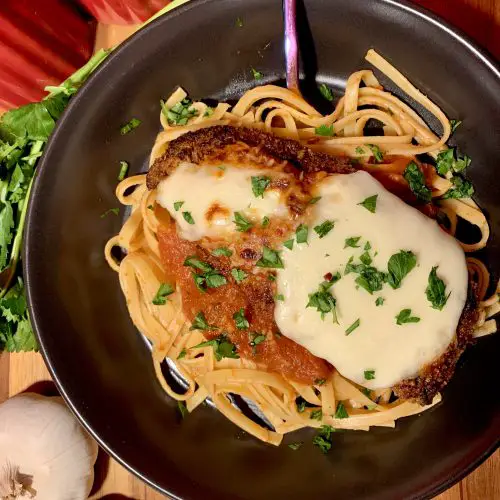 chicken parm main photo with black plate and surrounded by garlic and parsley