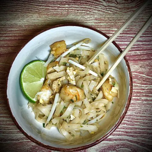 pad thai plated with chopsticks and served with lime wedge. rice noodles, scrambled eggs, bean sprouts, shrimp and chicken.
