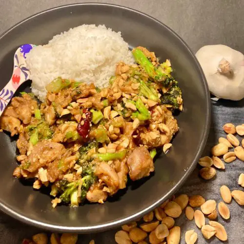 Thai peanut chicken with broccoli served with white rice. Served in black bowl with floral spoon and surrounded on cutting board by whole garlic and peanuts