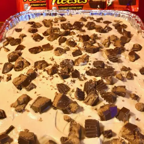 dark chocolate cake with peanut butter and cream cheese whipped frosting and topped with chopped Reese's cups. Pictured in a foil pan in front of red wall with three packs of Reese's cups propped up against it.