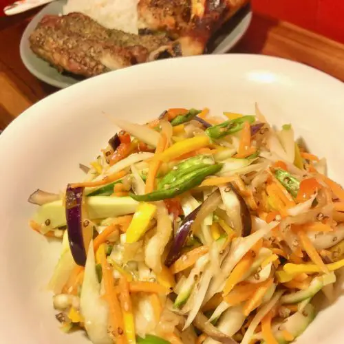 thai sum tom plated in white square bowl with a gray plate of jasmine rice, chicken drumsticks and ribs in the background. The gray plate has a floral spoon with the food