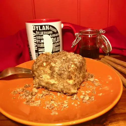 french toast casserole on orange plate surrounded by oats. pictured with a gold fork, a white coffee mug, jar of syrup and cinnamon sticks on a wooden table