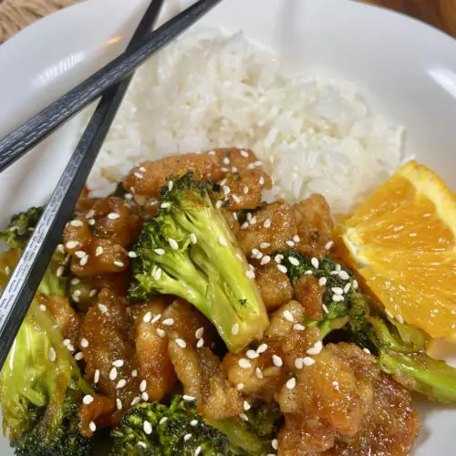 Orange Chicken with Broccoli pictures on a white square plate with Jasmine rice and a slice of orange. Black chopsticks are laying across the plate. There is a ramekin with fresh Thai chilis in the background.