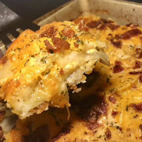 creamy potatoes au gratin with bacon. a corner piece is being held up out of the baking dish on a spatula so that you can see the layers of potato, onion and cheese