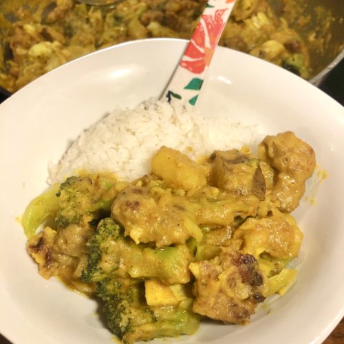 thai yellow curry with sausage and vegetables on white square bowl with rounded edges. photographed beside white rice with floral spoon. In the background is the large skillet with the rest of the dish.
