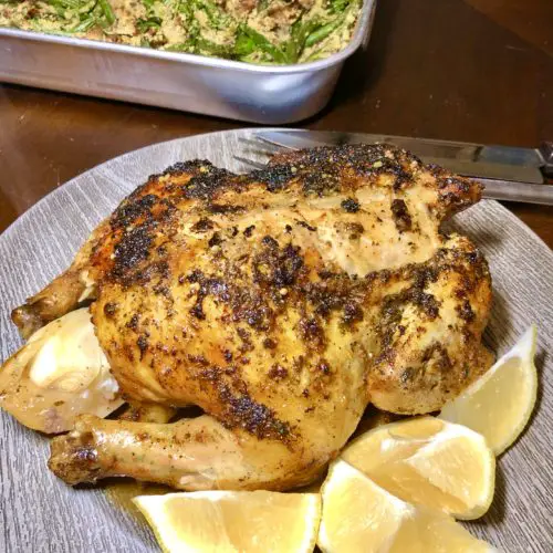 Lemony oven roasted chicken being served on gray plate with lemon wedges pictured with green bean casserole in the background