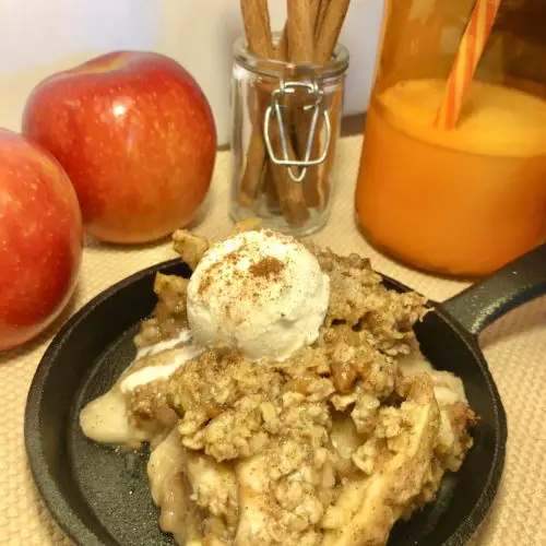 apple crisp topped with vanilla ice cream and a sprinkle of cinnamon in a mini cast iron skillet. skillet is photographed on tan fabric with two red apples, a jar with cinnamon sticks and an orange mason jar filled with milk in the background