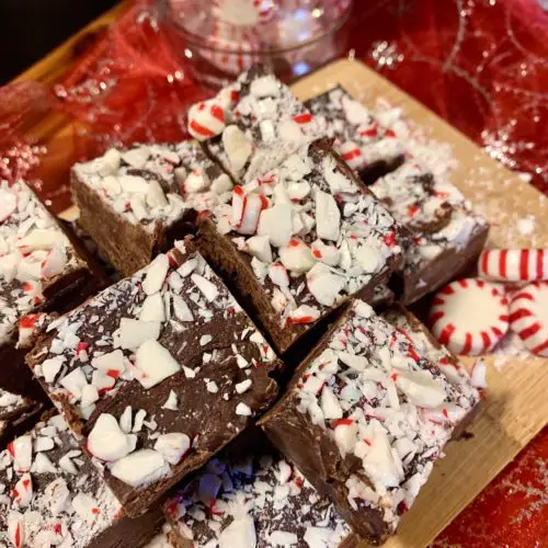 squares of peppermint fudge on oak cutting board pictured with starlight mint candies. These are on top of a red and silvery christmas fabric with a small jar filled with starlight mint peppermint candies