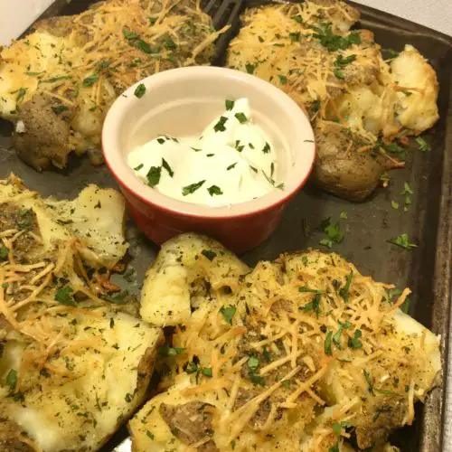 Four parmesan and olive oil smashed potatoes on a pan with a spatula under one potato and a red and white ramiken in the middle of the pan with sour cream. the entire pan and sour cream are garnished with fresh parsley