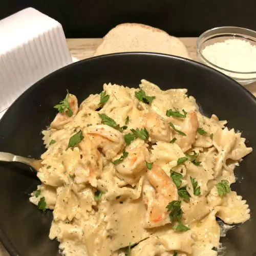 cajun shrimp pasta pictured in black matte bowl with fork in bowl. Pasta is garnished with fresh parsley. Bread, a butter dish and a glass ramiken with grated parmesan cheese are in the background