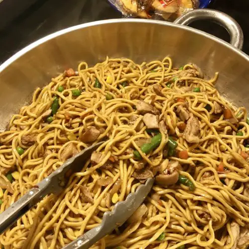 large silver skillet with chicken lo mein being tossed with tongs on stovetop. There is a blue bowl beside the skillet filled with Chinese fortune cookies