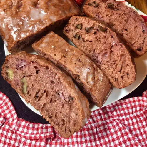 Four slices and a half loaf of strawberry walnut bread with lemon glaze on white square plate. Pictured with one strawberry and floral butter knife on top of wooden cutting board covered with white and red checkered fabric