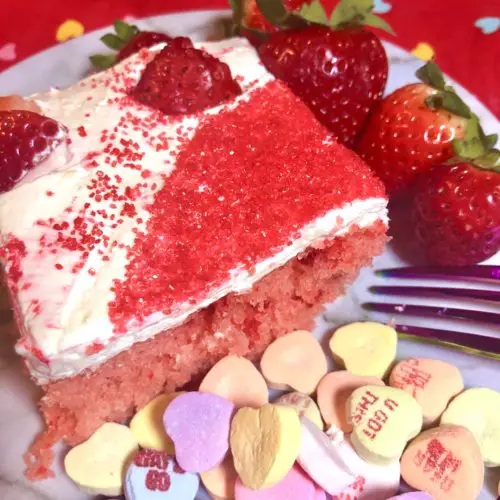 slice of strawberry cake with cream cheese and cool whip frosting. The cake is topped with fresh heart shaped strawberries and red sugar sprinkles. Played on white plate with purple fork. There are three fresh whole strawberries on the plate with scattered Sweetheart candies with words on them on the plate. There is red fabric with multi-colored hearts on the table in the background
