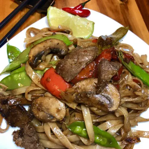 Thai beef rice noodles with mushrooms, snow peas and red bell peppers pictured on white square plate with lime wedge and two Thai chili peppers. Black chop sticks are pictured in the background and everything is on a wooden cutting board