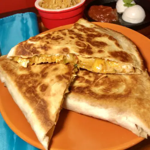 Three servings of cheesy chicken quesadillas with salsa arranged on an orange plate. There is a bright blue napkin pictured next to the plate and everything is on a wooden cutting board. In the background, there is a red ramekin with Spanish rice and beside that is a mini black cast iron skillet with salsa and sour cream for dipping.