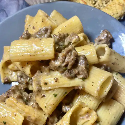 Homemade Hamburger Helper made with rigatoni pasta and a cheese sauce with ground beef. Pasta is plated on a gray plate with a white ramekin of grated parmesan cheese and a round slice of garlic bread in the background.