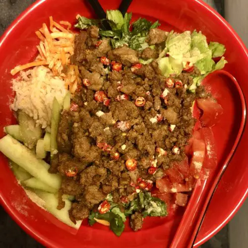 Thai caramelized ground pork in a Jasmine rice bowl. The rice bowl contains lettuce, cucumber, carrots, cilantro, mint, Thai chilis and toasted rice powder. Pictured in a red bowl with a red soup spoon and black chopsticks on top of a charcoal marbled table.