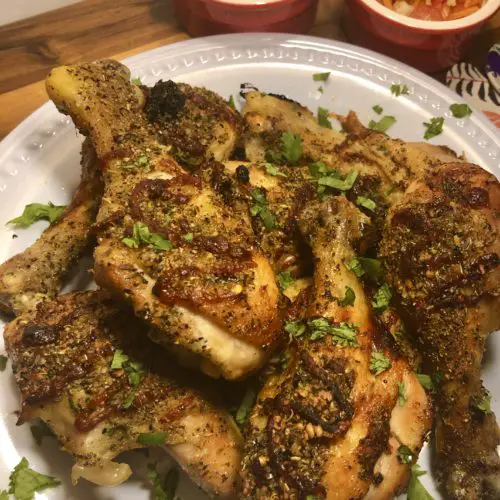 Baked honey sriracha chicken drumsticks plated on a white plate and garnished with chopped cilantro. The plate is on a wooden table with two ramekins in the background. One ramekin has Jasmine rice and one has som tum (Thai carrot salad).