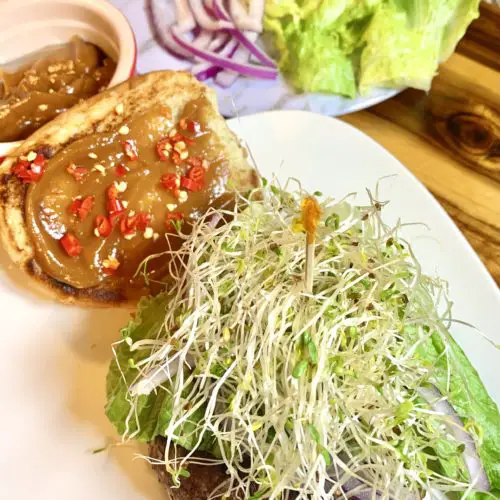 Thai pork burger on white square plate and topped with bok choy, sprouts and purple onion. The top bun is beside the burger on the plate and topped with peanut sauce and thinly sliced Thai chili peppers. There is a red ramiken with additional peanut sauce behind the plate. There is a white marble plate with additional vegetables for topping. Everything is pictured on a wooden cutting board.