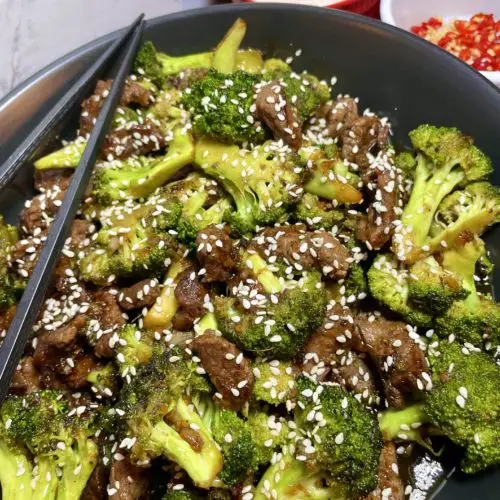 Better Than The Buffet Beef and Broccoli pictured in a black bowl with black chopsticks across the top of the bowl. The dish is garnished with sesame seeds. There are ramekins in the background. One has Jasmine rice and one has fresh Thai chili peppers that have been sliced.