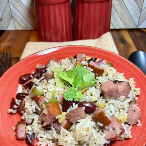 Red Beans and Rice pictured with a cilantro garnish on top. The red beans and rice is on a red plate over a tan cloth on a wooden cutting board. There are oversized red salt and pepper shakers in the background and matte black fork and spoon beside the red plate.