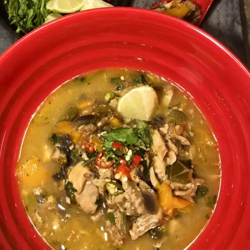 Moqueca (Brazilian Fish Stew) plated in red bowl and garnished with cilantro and fresh Thai chilis. There is a mini cast iron skillet in the background with lime wedges, cilantro and Thai chili peppers. There is a red ramekin in the background with white rice. Everything is on top of a dark gray marbled surface.