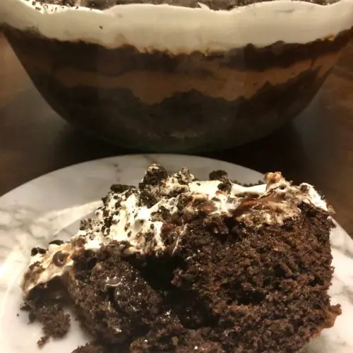 A serving of Chocolate Dirt Cake (Mud Bucket) on a white marbled plate. A trifle dish is behind the plate showing layers of chocolate and Cool Whip.