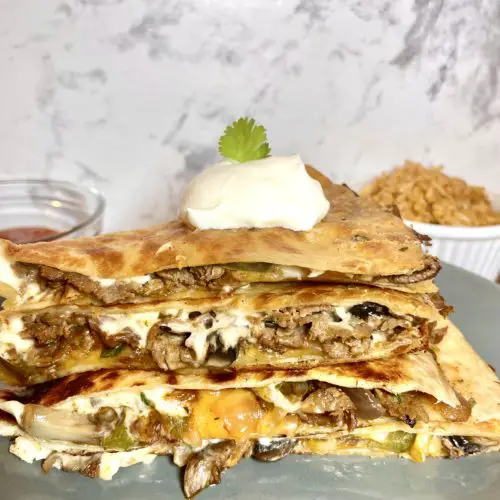 A stack of four steak bomb quesadilla pieces on a gray plate. A dollop of sour cream and a sprig of cilantro are on the top of the stack. There is a white ramekin in the background with Spanish rice and a red ramekin with salsa.