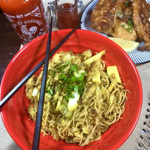 Singapore noodles in a red bowl with black chopsticks lying across the top of the bowl. Sriracha, a jar of chili sauce and a small plate of Chinese chicken fingers are behind the bowl.