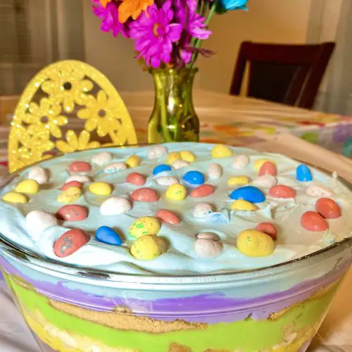 The Best Easter Trifle Dessert pictured showing bright colorful layers of cake, vanilla pudding, funfetti morsels, cream cheese and Cool Whip. The trifle is topped with blue dyed Cool Whip and Whoppers Mini Robin Eggs.